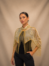 A woman modeling a gold and beige embroidered fringed bolero jacket over a black top and black pants, set against a neutral beige studio backdrop.