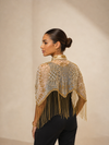 A woman shown from the back wearing a sheer beige and gold embroidered shawl with long fringe, paired with black pants, against a neutral beige studio background.