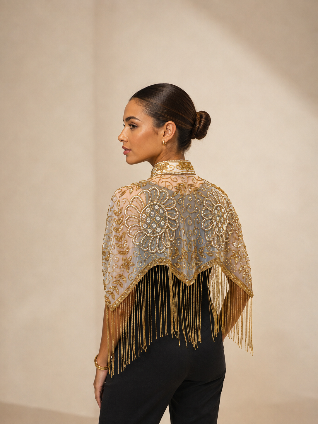 A woman shown from the back wearing a sheer beige and gold embroidered shawl with long fringe, paired with black pants, against a neutral beige studio background.